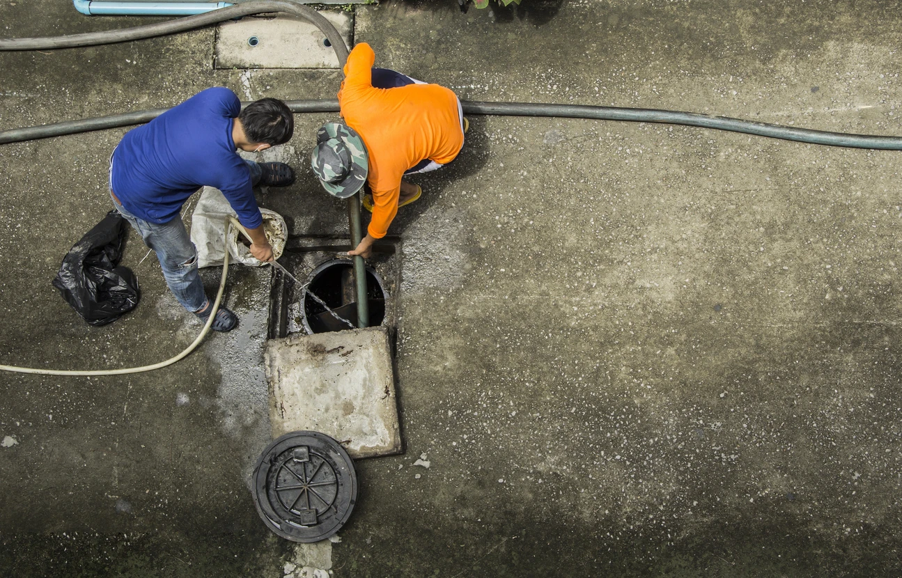 Deux techniciens en combinaisons bleue et orange travaillent sur un regard d'assainissement en béton