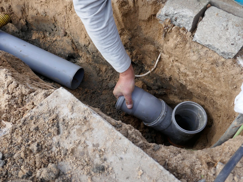 Scène extérieure avec puits en béton circulaire muni d'un couvercle et d'une poignée métallique, situé dans un terrain herbeux avec végétation éparse et pierres dispersées