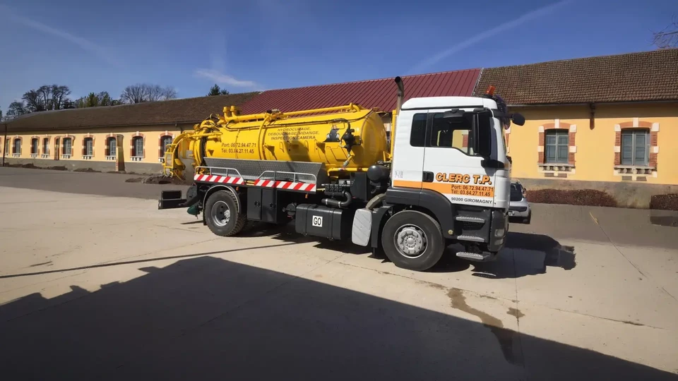 Camion d'assainissement jaune stationn&eacute; devant un b&acirc;timent historique en pierre, tuyaux verts d&eacute;ploy&eacute;s vers une bouche d'&eacute;gout pour vidange ou curage